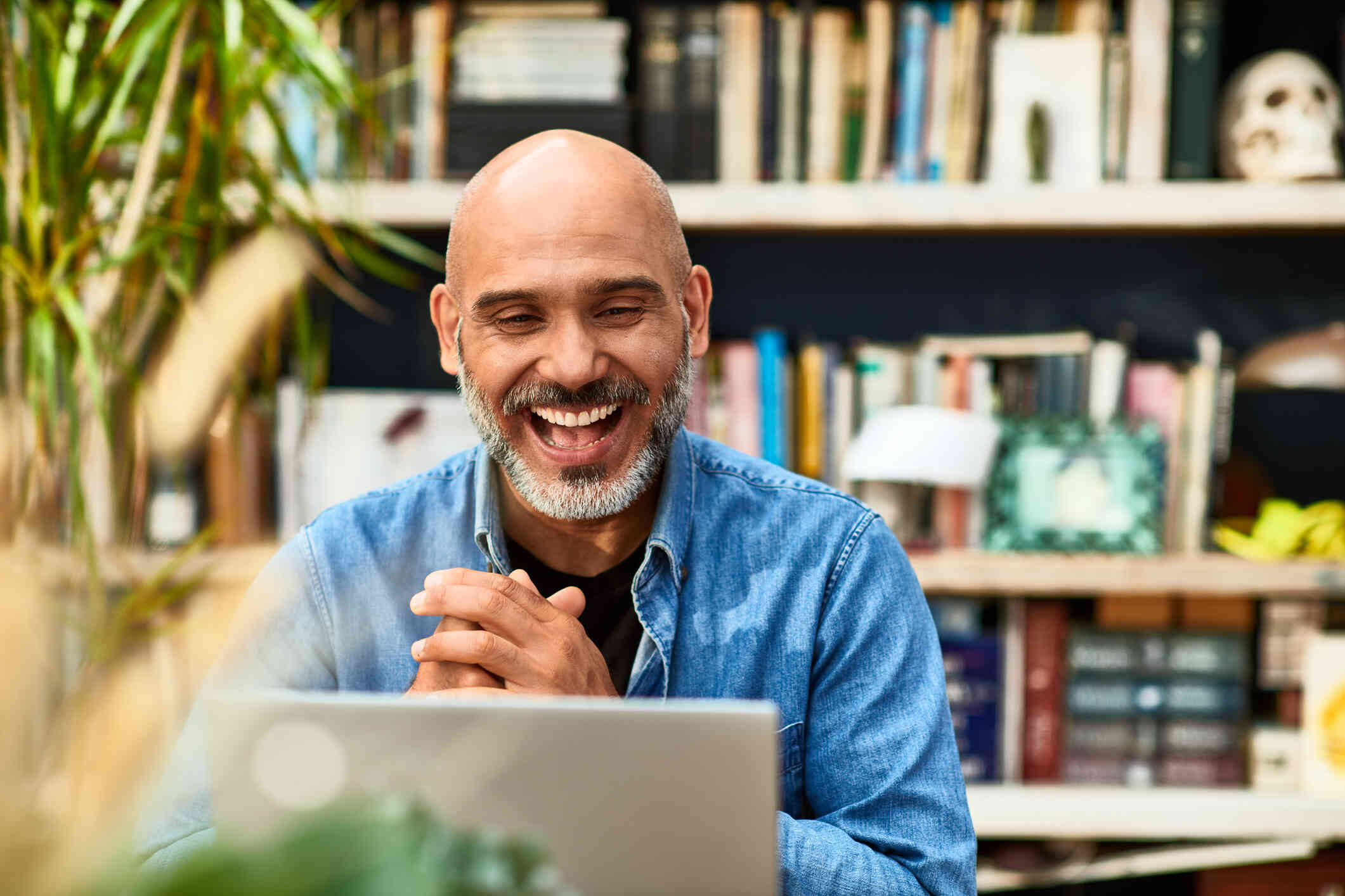 A middle aged man sits at a table and smiles with his hands clasped together while looking at the laptop infront of him.