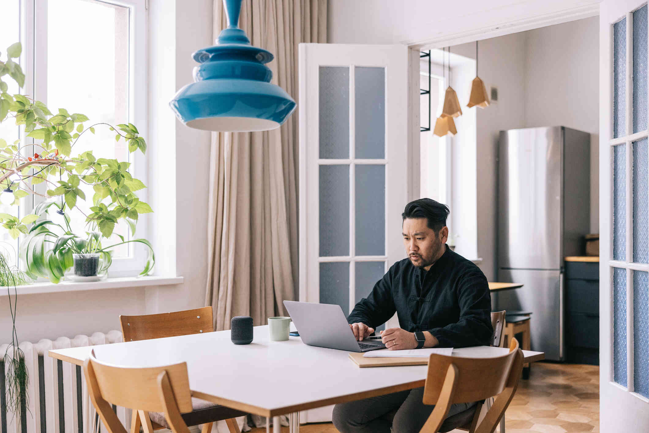 A man in a black shirt sits at his kitchen table and looks at the laptop open on the table infront of him with a serious expression.